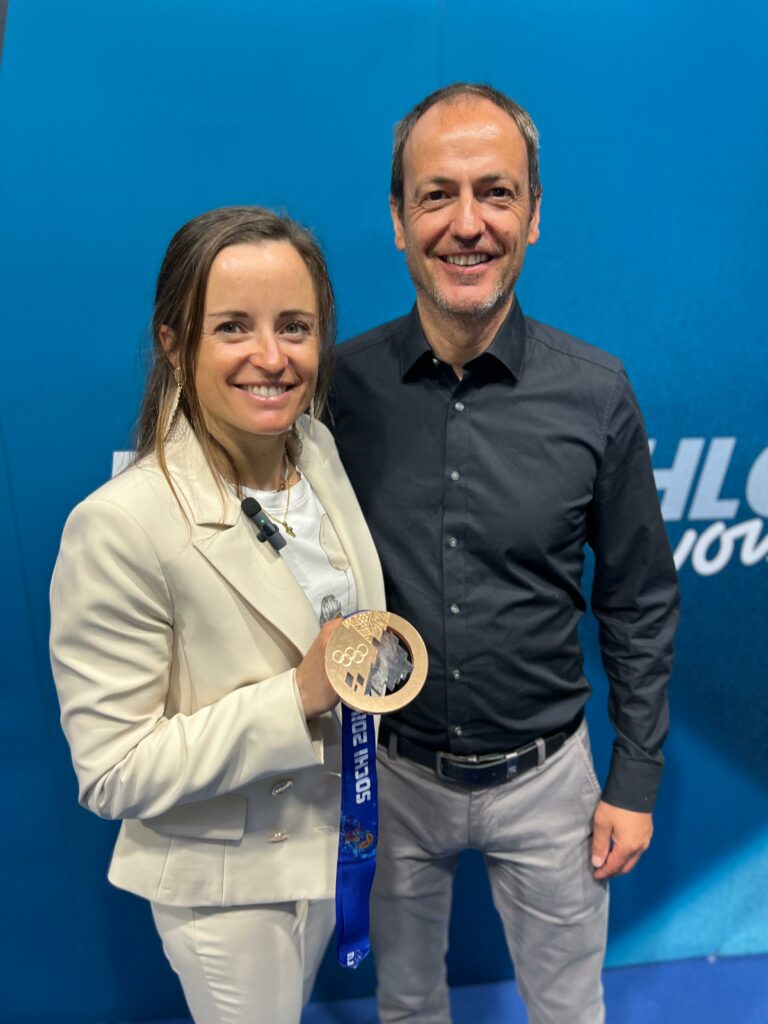 Eine Frau in hellem Hosenanzug hält eine olympische Bronzemedaille mit blauem Band in der Hand. Neben ihr steht ein Mann in dunklem Hemd. Beide lächeln in die Kamera vor einem blauen Hintergrund.