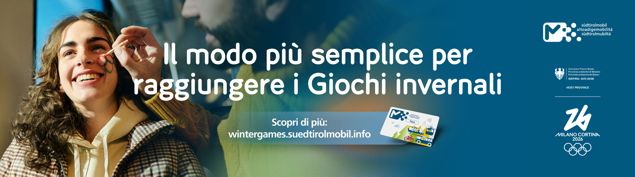 Giovane donna sorridente mentre le vengono dipinti gli anelli olimpici sul viso. Testo nell’immagine: ‘Il modo più semplice per raggiungere i Giochi Invernali’, con riferimento a wintergames.suedtirolmobil.info e i loghi di altoadigemobilità, Provincia autonoma di Bolzano e Milano Cortina 2026.